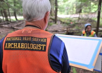Un hombre que lleva puesto un chaleco de seguridad anaranjado con la inscripción "National Park Service Archaeologist" en la espalda mira un tablero en el que hay un papel con información topográfica.