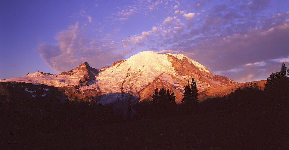 Una gran montaña cubierta de glaciares resplandece en tintes rosas y rojos al amanecer.