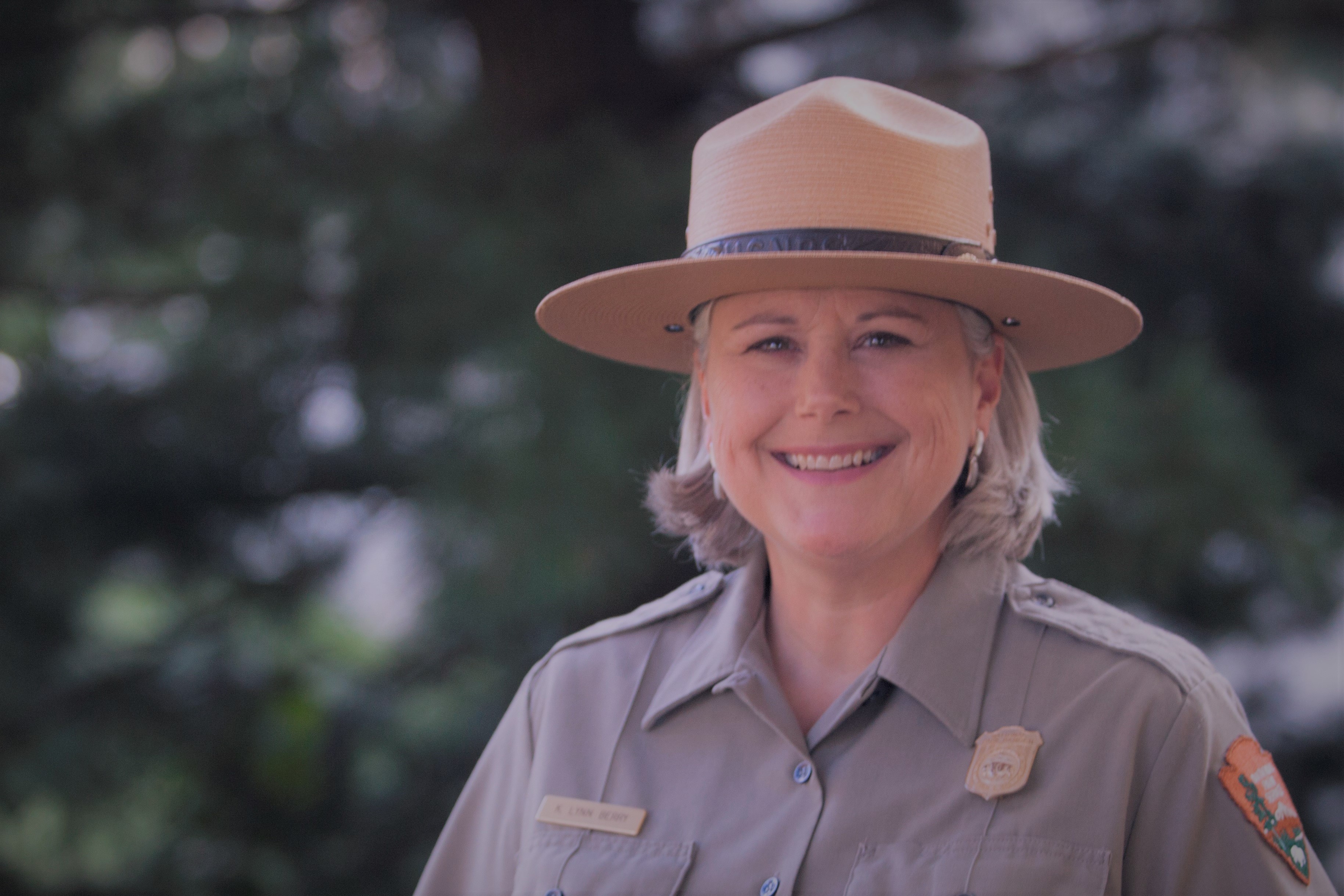 Photograph of older middle age woman in ranger uniform and hat, smiling at camera.