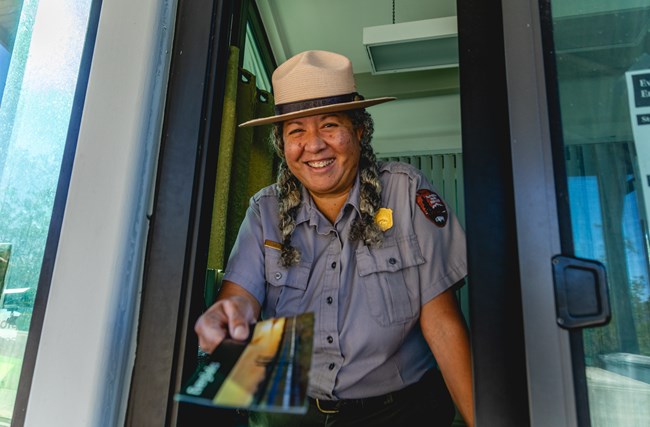A park ranger stands in the entrance station booth while smiling and holding an Everglades pamphlet.