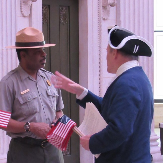 In Federal Hall a park ranger is speaking with a person dressed in colonial attire, holding small American flags.