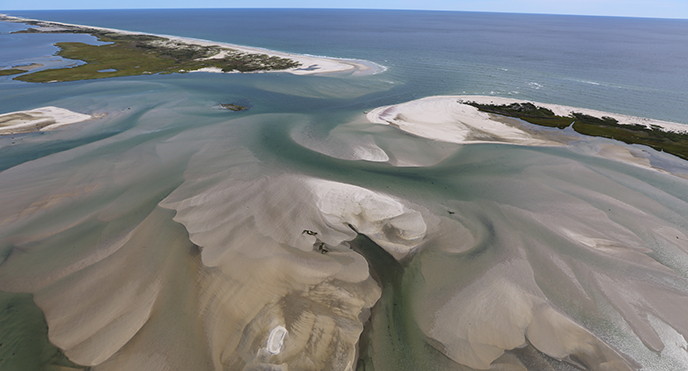 Looking down from the sky, sand swirls just below the ocean surface around sandy islands with light vegetation.
