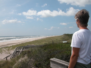 Woman looks over dunes and beach to rough ocean surf.