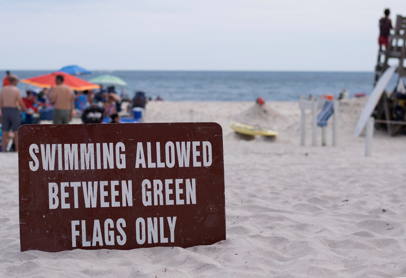 A sign reading, "Swimming allowed between green flags only" sits in the sand near a lifeguarded beach.