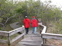 Two red-coated walkers stroll down wooden boardwalk into the forest.