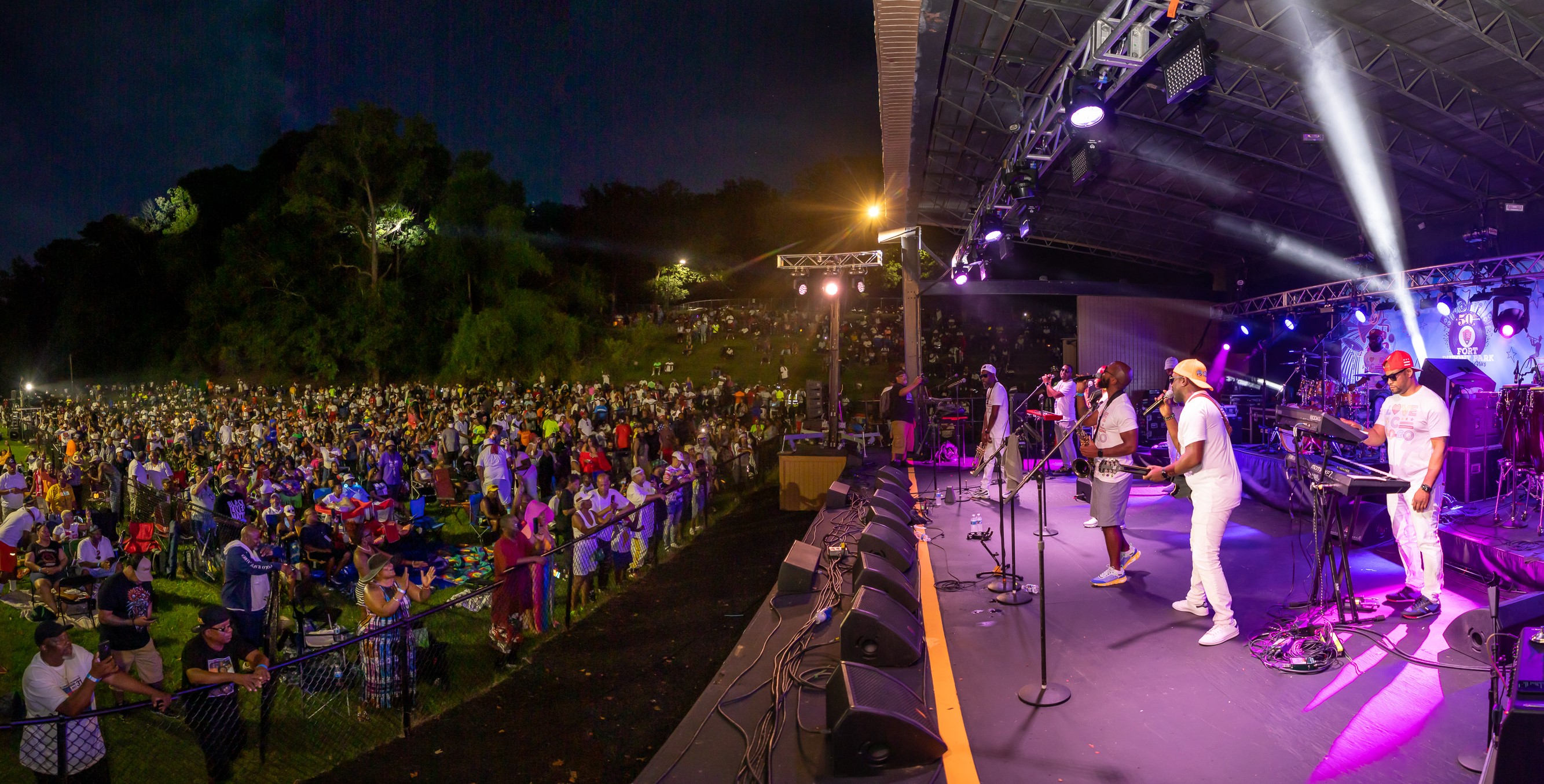 A band wearing white shirts bathed in purple light performs on stage for a crowd on a lawn at night.