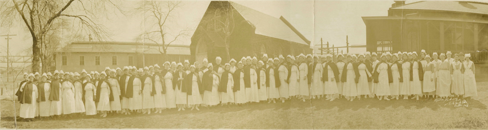 A black and white image of a large group of nurses posing in front of hospital buildings being constructed in the background.