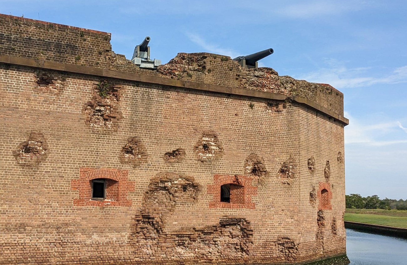 A masonry wall of a fort with openings for cannon and a variety of damage and battle scars. Two cannon poke over the top of the upper walls. They are further exposed by the wall that had been torn away during a battle.