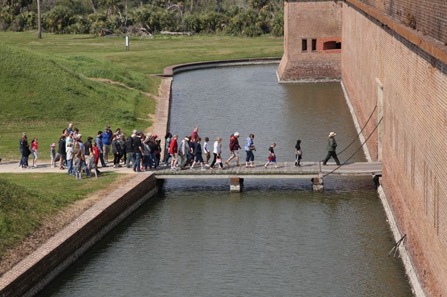 A park ranger leads a large group of people over a wooden bridge across the moat.