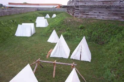 a lush green grassy hole is filled with large, white, triangular, canvas tents. wooden walls and shadows loom over them.