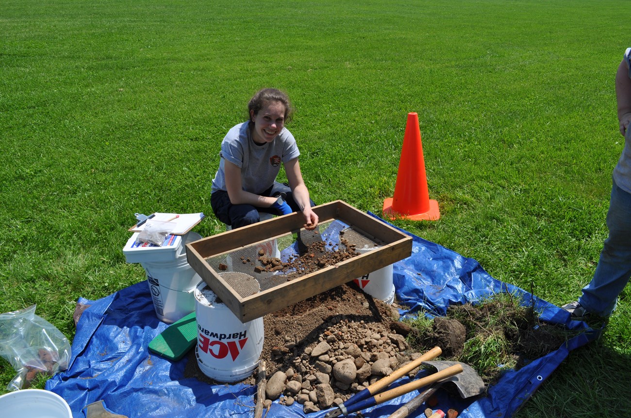 A woman sits in the grass with a hole dug in front of her, surrounded by archeological equipment like sivs and trowles.