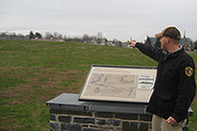 a man points over a grassy green field under a grey sky to the fort