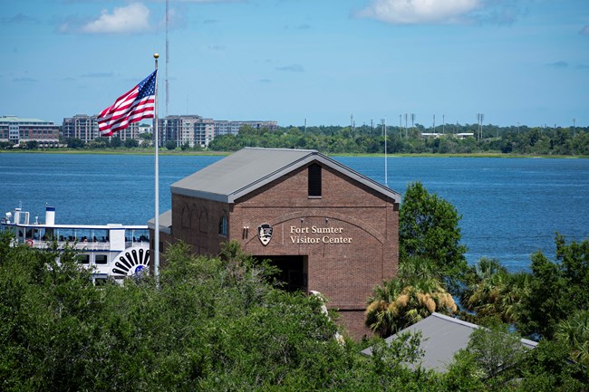 A brick building surrounded by trees, a boat in the backround and the US flag flying.