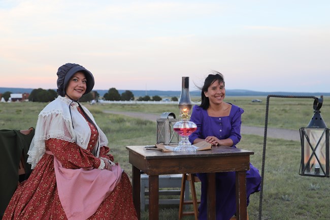 Two women sitting at a table with an oil lamp, both wearing long 19th centur dresses