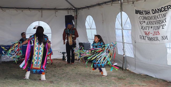 Two Navajo dancers wearing colorful costumes twirl around while a man beats a drum in background.