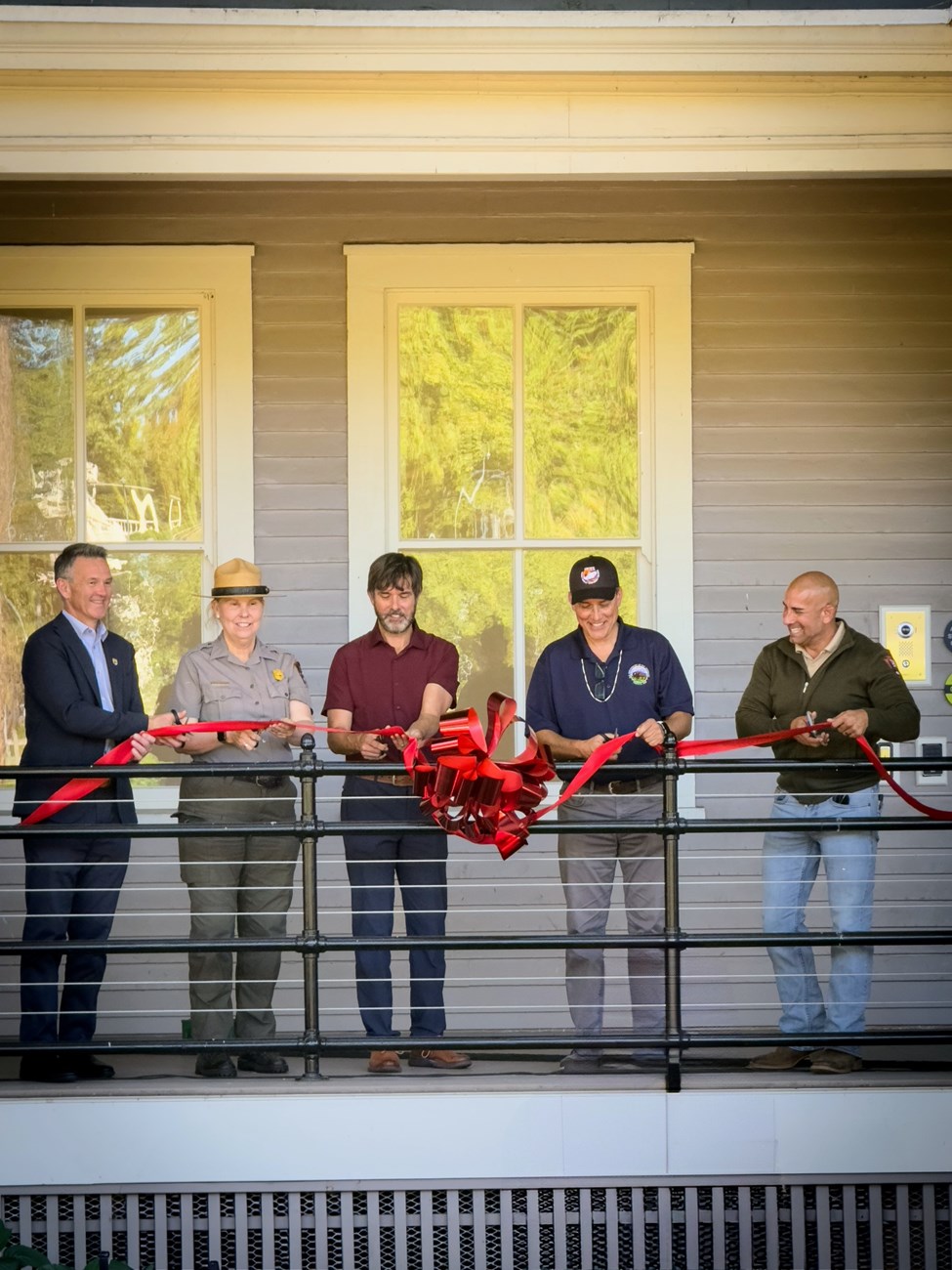 Multiple people stand on the porch of a grey building holding a red ribbon and scissors, preparing to cut the ribbon to officially open the building. 