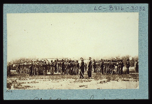 A black and white photograph of African American civil war soldiers standing in rows. A few white soldiers/officers stand to the side.