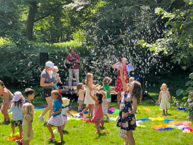 Group of children play on lawn with bubbles while person sings on stage