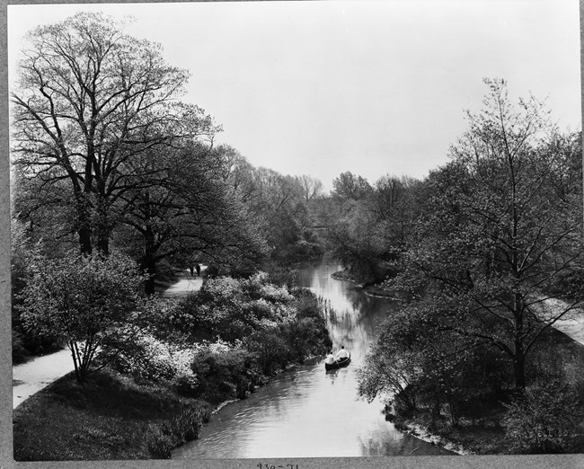 Black and white of body of water with people on canoe in middle with paths lined with trees on both sides.