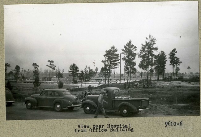 Black and white photograph of large dirt area with trees lined up. In front of the dirt are 3 cars.