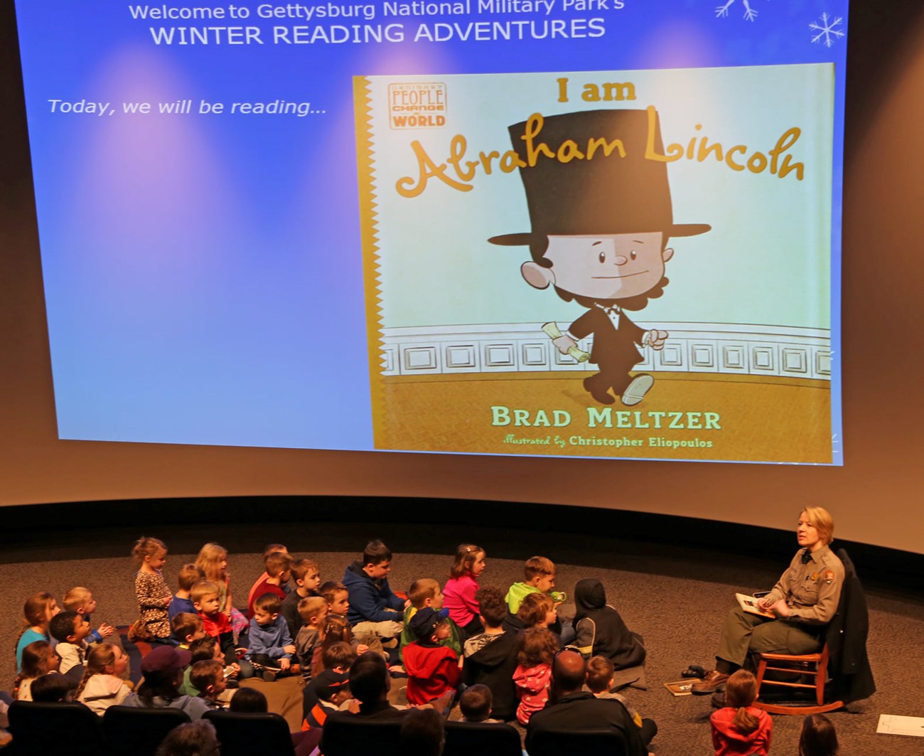 A Park Ranger reads to a group of small children. On the screen is the cover of the book I am Abraham Lincoln