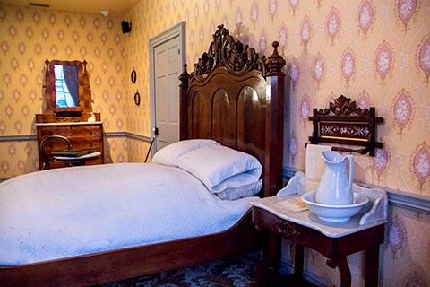 A bed with a large, ornate wooden head board sits next to the wall. On the near side is small table with a white marble top and white pitcher. On the far side is a small dresser with a mirror.