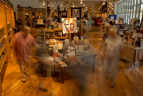 Visitors move around the Museum Bookstore. There are multiple people looking at book and walking around tables stacked with books.