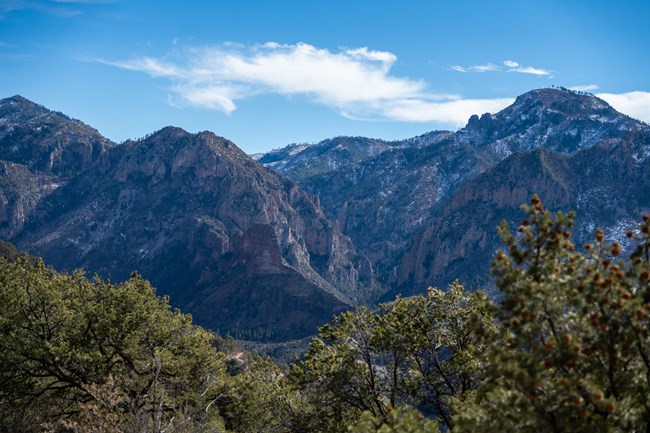 A view of a deep canyon with towering mountains on the opposite side, some of the slopes have snow and the sky is blue.