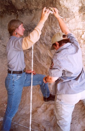 A photo of archeologists measuring the side of a cave wall.
