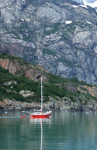 A red and white sailboat tows a dinghy in calm reflective water with rocky cliffs behind