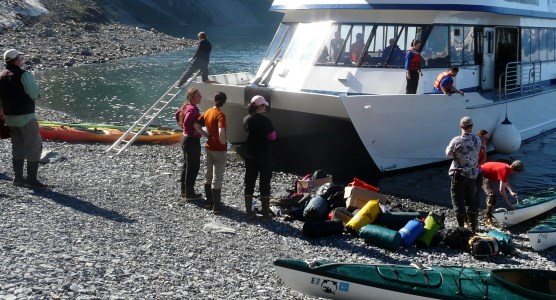 Campers standing on shore with the catamaran-style "day-boat" unloading kayaks and other gear for the campers to use.