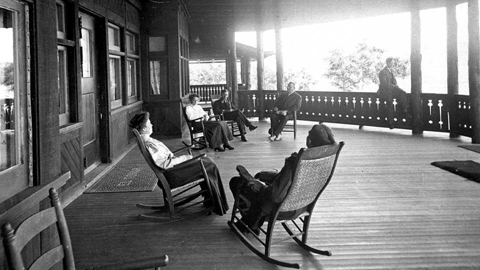 Black and white historic photo of El Tovar Hotel front porch with several well dressed people reclining in rocking chairs