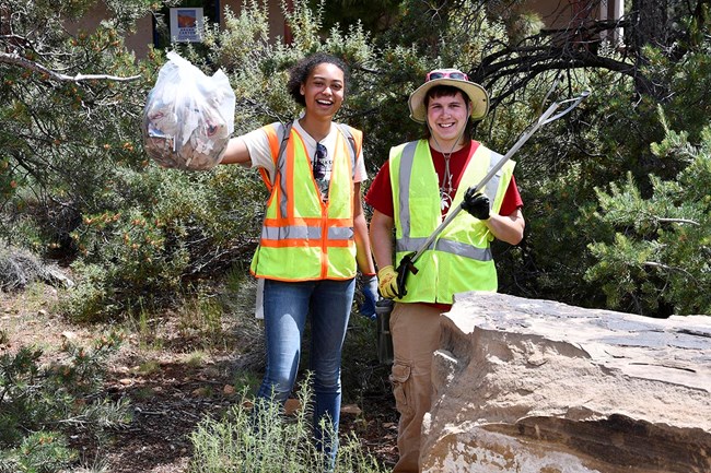 Two college students, wearing safety vests are posing for a photo with a trash picker and a bag full of trash.