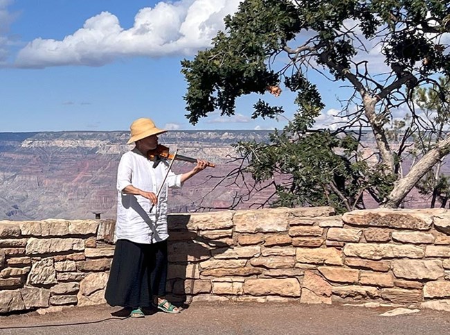 A woman wearing a white shirt and a large sun bonnet is playing a violin near a stone guard wall overlooking the Grand Canyon landscape.