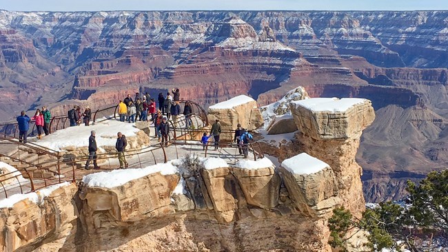 a scenic overlook on top of a large rock outcropping. Snow covers the ground. Small groups of people are behind metal guardrails and viewing a vast canyon landscape of colorful peaks and cliffs.