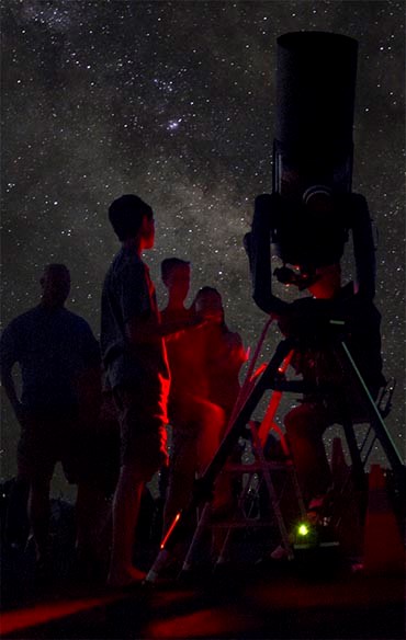a starry night. a family is gathered around a large telescope as an astronomer is talking. Everyone is bathed in red light.