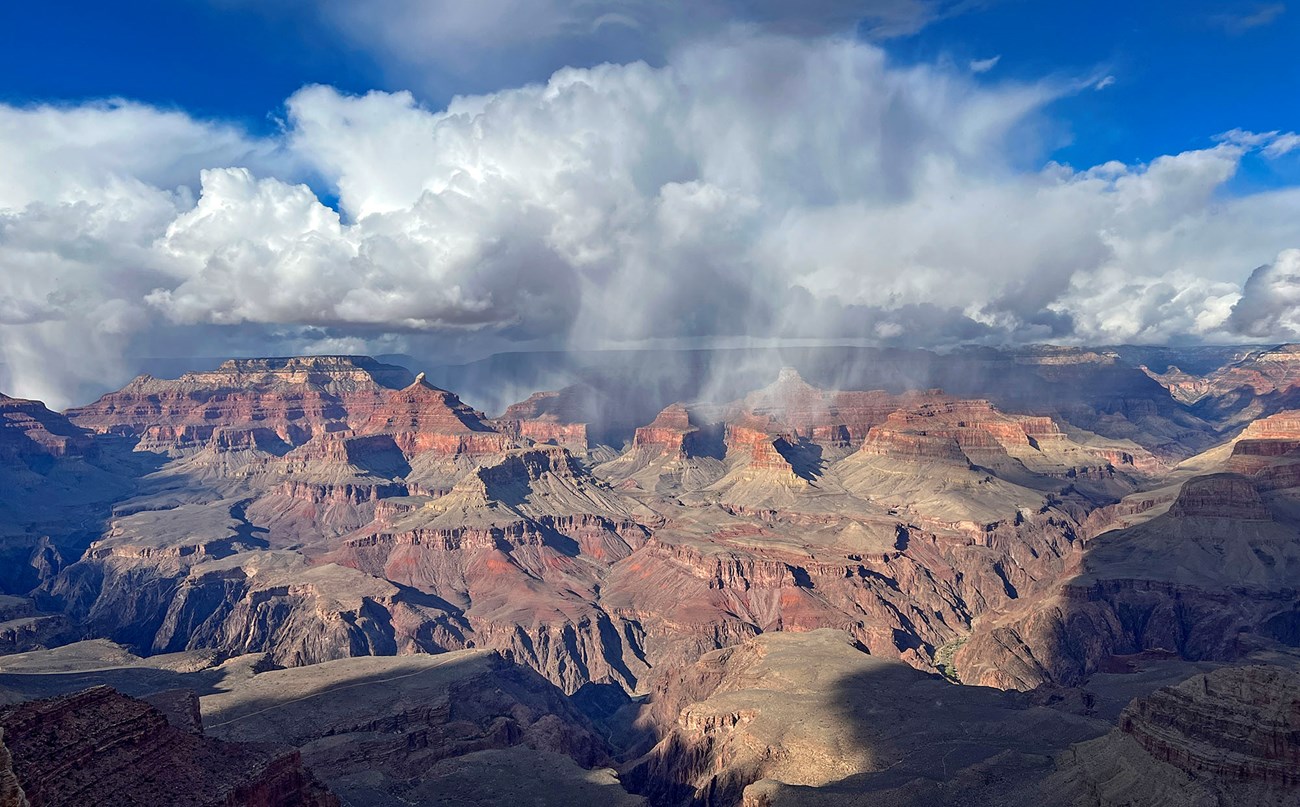 With a bright blue sky in the background, a mass of winter storm clouds is slowly rising and mingling between the vermilion-colored peaks and cliffs within the vastness of Grand Canyon.