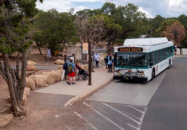 Passengers about to board a white and green bus in a forested area with converging footpaths. Bus marque reads, Orange Route.