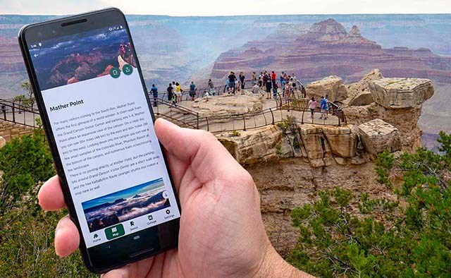 a hand holding cellular phone that displays photos and text for a location. In the background, people at a scenic overlook with peak visible in the distance