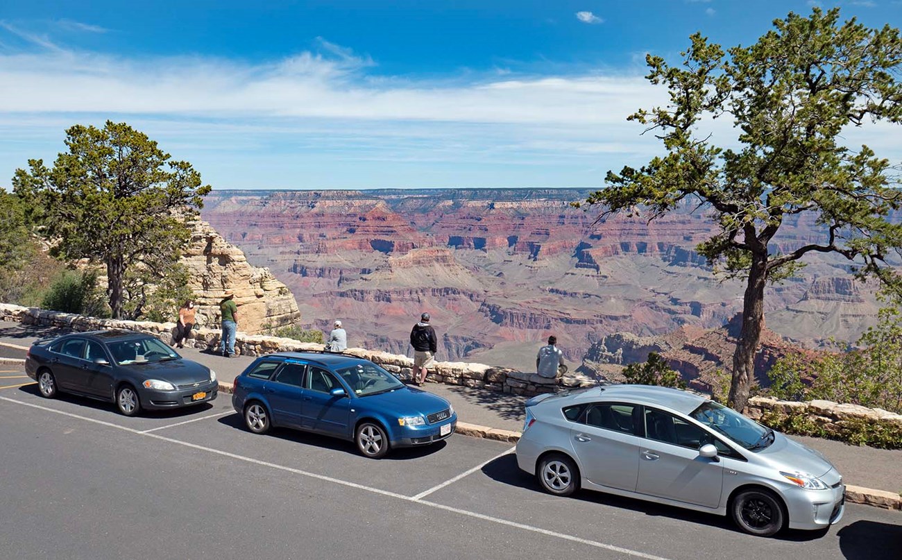 three cars parked at a scenic overlook with five people standing beside a rustic stone guard wall on the edge of a vast and colorful canyon,