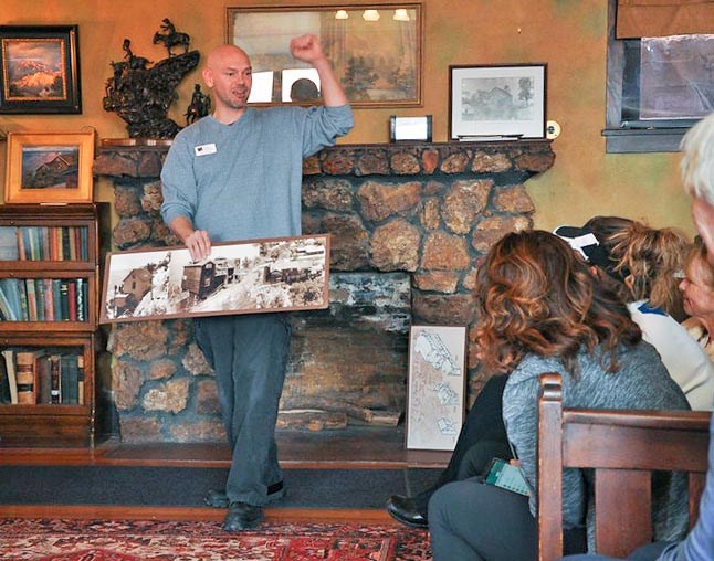 A tour guide is standing in the parlor of an historic home and holding up three photos that show the stages of the building's expansion over the years. Several people are sitting in chairs and listening to his presentation