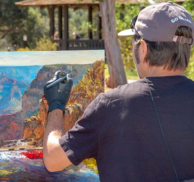 An artist wearing a t-shirt and cap is painting a landscape of canyon peaks and cliffs.