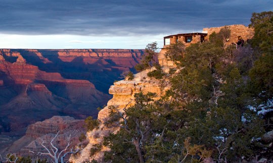 A rustic stone building on the edge of a cliff that overlooks colorful cliffs and peaks at sunset.