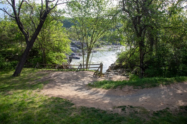 A gravel trail approaches a wooden fence at the lip of a steep rocky trail with a river visible down below.