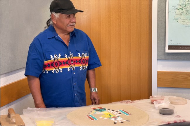 Mitchell Silas stands speaking over a sand painting depicting an abstract figure in turquoise, blue, yellow, black and white