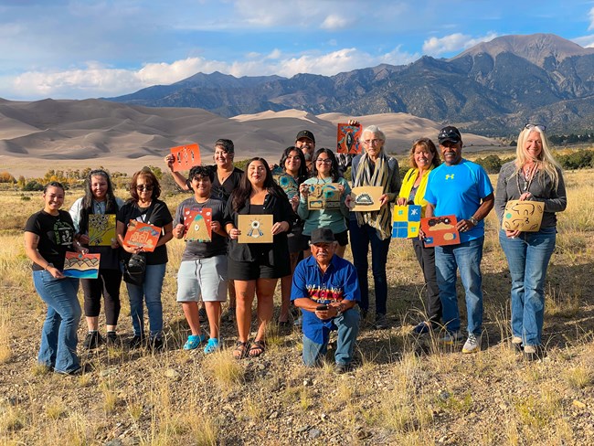 Navajo sand painter Mitchell Silas kneels in front of a group of participants holding sand paintings. In the background are dunes and mountains.