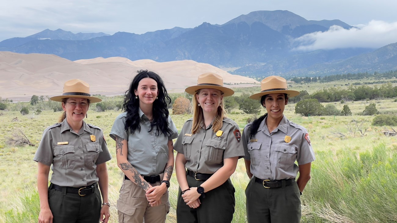 Four women in uniform standing shoulder to shoulder with sangre de cristos and dunes in the background