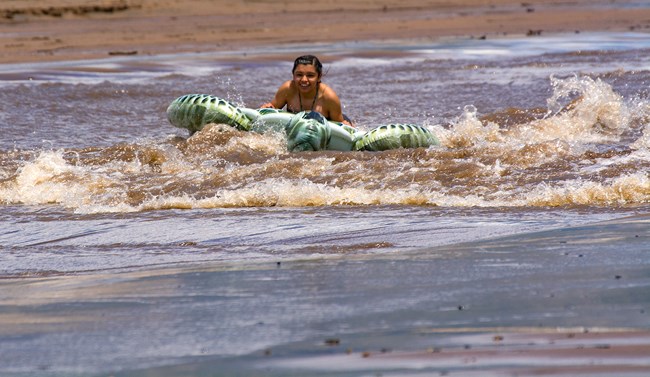 A teen girl rides a turtle-shaped floatation toy on a large wave flowing over sand