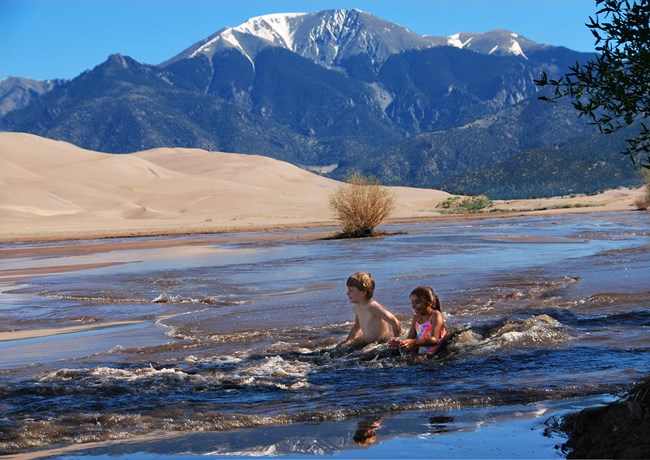 Two children sit in a rushing wave in Medano Creek, flowing at the base of dunes and a mountain