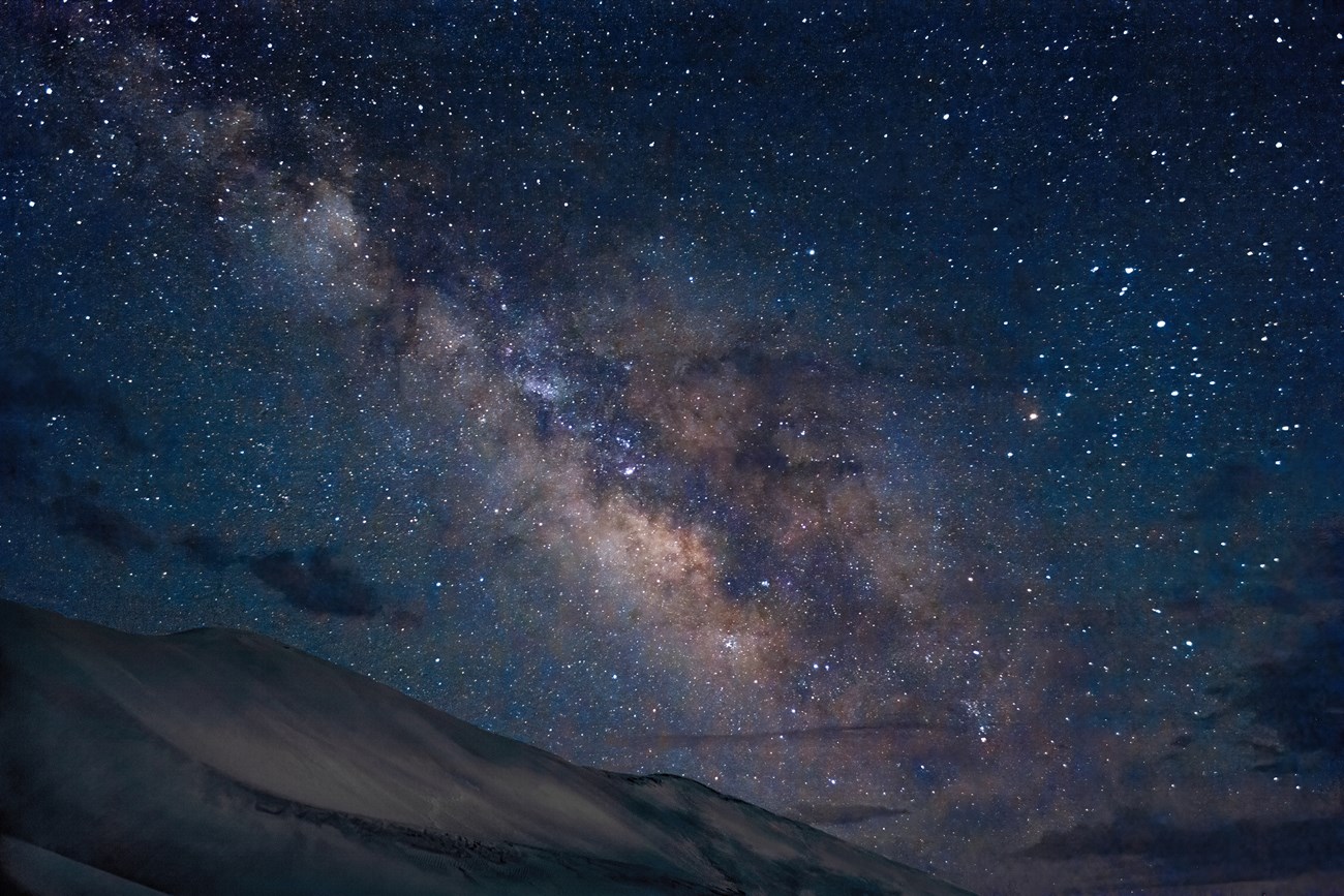 The Milky Way appears as a soft, linear cloud among stars over a dune ridge.